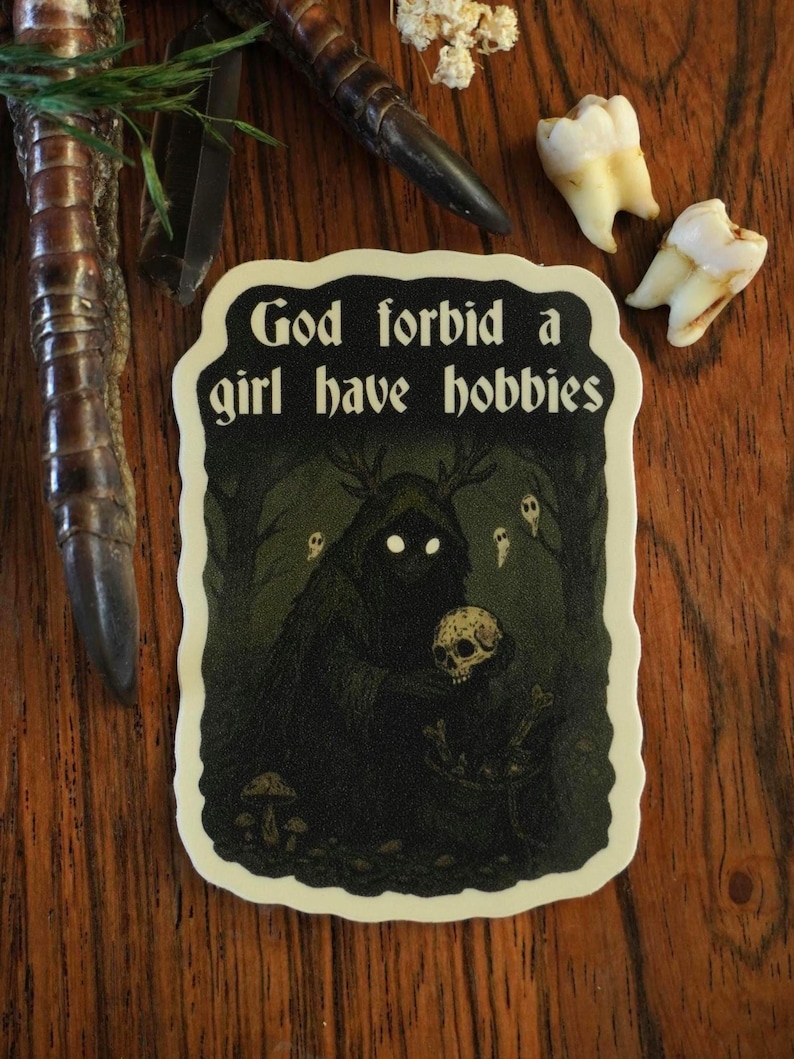 Może przedstawiać: Naklejka z napisem "God forbid a girl have hobbies" w czarno-białym wzorze. Naklejka przedstawia mroczną scenę leśną z stworzeniem trzymającym czaszkę. Dodatkowe elementy to zęby i dekoracyjny pazur.