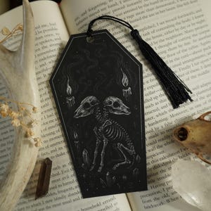 May include: Black coffin-shaped bookmark with a detailed illustration of a two-headed skeletal creature, candles, and crystals. The bookmark has a black tassel and is set against an open book.