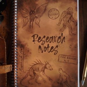 May include: Spiral-bound notebook with a brown cover featuring sketches of cryptids. The cover reads "Research Notes" with illustrations of a moth-like creature, Bigfoot, and other unknown creatures. Includes stamps for Point Pleasant, W.V. and Pine Barrens, N.J.