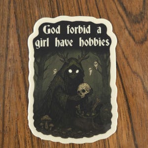 Może przedstawiać: Naklejka z napisem "God forbid a girl have hobbies". Ilustracja przedstawia mroczną postać z rogami trzymającą czaszkę, otoczoną lasem i duchami. Naklejka ma białą obw&oacute;dkę.
