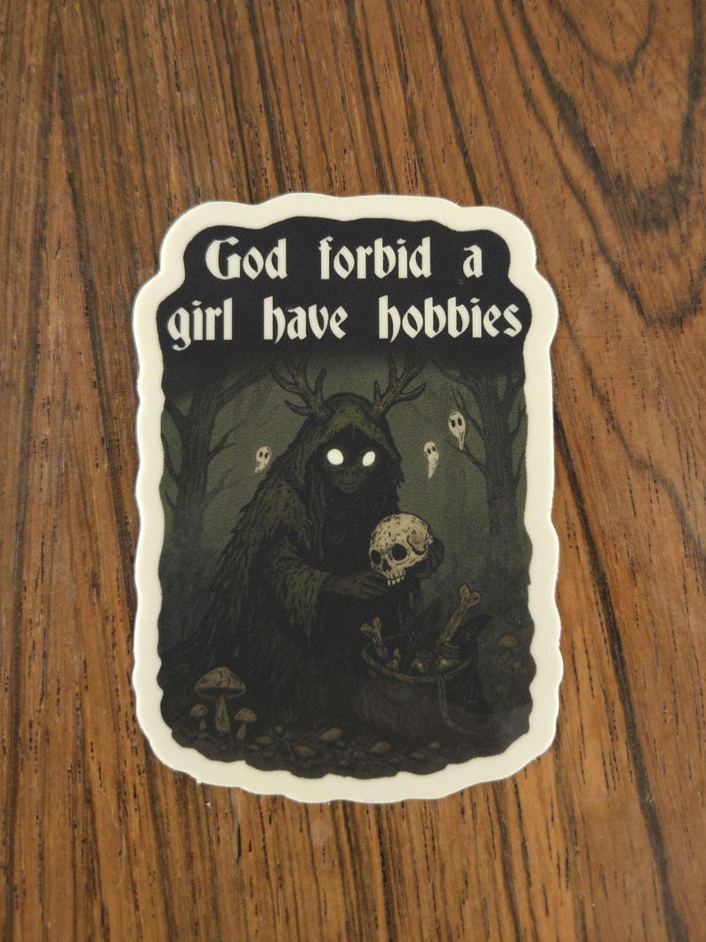 Może przedstawiać: Naklejka z napisem "God forbid a girl have hobbies". Ilustracja przedstawia mroczną postać z rogami trzymającą czaszkę, otoczoną lasem i duchami. Naklejka ma białą obw&oacute;dkę.
