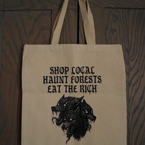 May include: Beige canvas tote bag with the text "SHOP LOCAL HAUNT FORESTS EAT THE RICH" in black gothic font. Below the text is a black illustration of a three-headed wolf. The bag has beige handles.