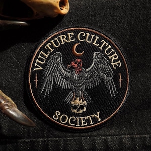 May include: Black embroidered patch with the words "VULTURE CULTURE SOCIETY" surrounding a vulture with outstretched wings, a crescent moon, and a skull. The patch is circular with a brown border.