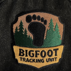 May include: Embroidered patch with a black Bigfoot footprint, green pine trees, and the text "BIGFOOT TRACKING UNIT" on a brown and gold background. The patch is sewn onto a black denim fabric.