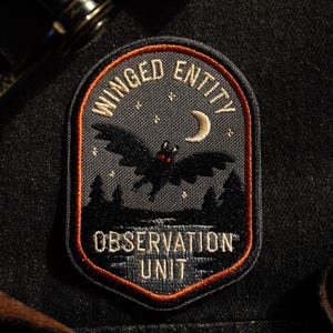May include: Embroidered patch with the words "Winged Entity Observation Unit." The patch features a black bat with red eyes flying in front of a crescent moon and a forest silhouette, all on a dark gray background.
