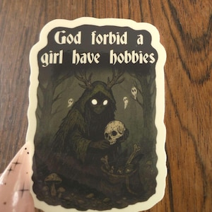 Może przedstawiać: Naklejka z napisem "God forbid a girl have hobbies". Ilustracja przedstawia postać w kapturze z rogami trzymającą czaszkę, otoczoną lasem i duchami. Paleta kolor&oacute;w to ciemna zieleń, czerń i biel.