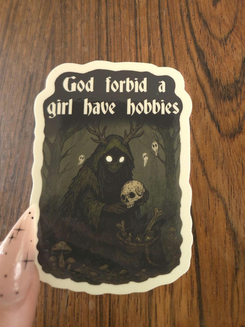 Może przedstawiać: Naklejka z napisem "God forbid a girl have hobbies". Ilustracja przedstawia postać w kapturze z rogami trzymającą czaszkę, otoczoną lasem i duchami. Paleta kolor&oacute;w to ciemna zieleń, czerń i biel.