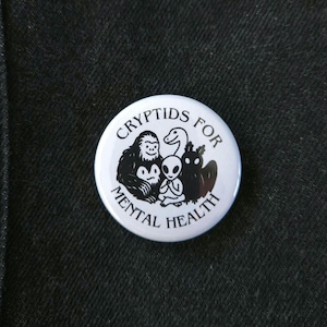 May include: White button with black text and illustrations. The text reads "Cryptids for Mental Health". The illustrations are of a Bigfoot, a dragon, an alien, and a Nessie.