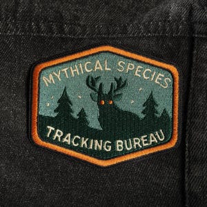 May include: A rectangular patch with the text "MYTHICAL SPECIES TRACKING BUREAU". The patch features a dark green silhouette of a deer with glowing eyes, set against a teal background with trees and stars, framed by an orange border.