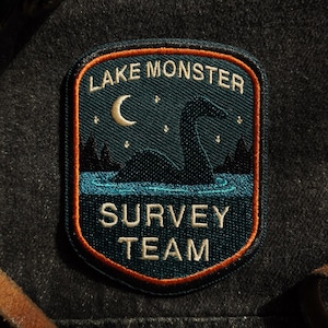 May include: Embroidered patch with the text "LAKE MONSTER SURVEY TEAM." The patch features a dark blue lake scene with a silhouette of a creature, a crescent moon, and stars. The patch has an orange border.