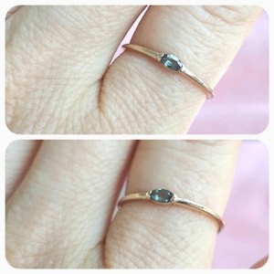 Puede incluir: Un delicado anillo de oro con una pequeña piedra preciosa ovalada de color verde oscuro. El anillo se lleva en un dedo.