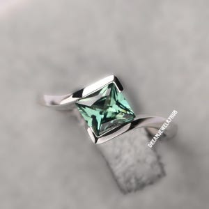May include: A silver ring featuring a square, emerald-cut, light green gemstone. The band has a modern, flowing design. The ring is set against a soft, neutral background. The text "DREAMJEWELRYHUB" is visible on the ring.