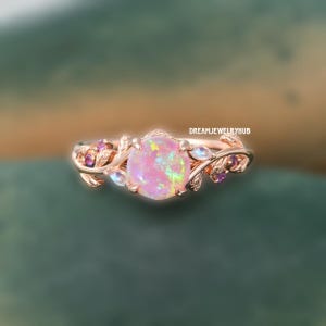 Puede incluir: Anillo de oro rosa con una gran gema de ópalo rosa redonda. El anillo está adornado con detalles de hojas y vides, junto con piedras preciosas rosas y azules más pequeñas. El texto "DREAMJEWELRYHUB" es visible.