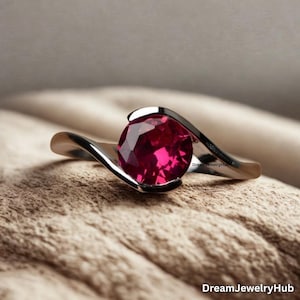 Può includere: Un anello d'argento con una grande gemma di rubino rotonda e sfaccettata. La fascia dell'anello ha un design fluido e asimmetrico che si curva attorno al rubino. La gemma è di un rosso vibrante.