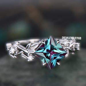 Könnte beinhalten: Ein silberner Ring mit einem quadratischen, schillernden Edelstein, der in der Mitte sitzt. Die Ringschiene ist mit zarten, blattartigen Details und kleinen, klaren Edelsteinen verziert.