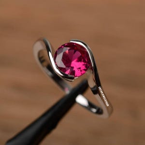 Peut inclure: Une bague en argent avec une pierre précieuse rouge vif sertie dans un design en spirale. La bague est sur un présentoir noir.