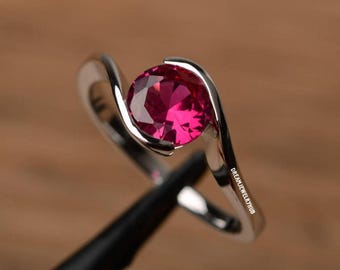 May include: A silver-coloured ring featuring a round, faceted, vibrant pink gemstone set in a bypass-style band. The metal band curves around the stone, and the text DREAMJEWELRYHUB is printed in small white letters along the side of the ring shank.