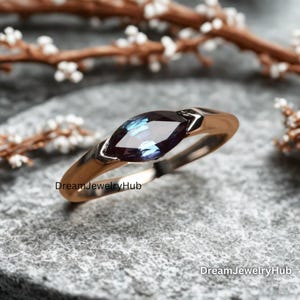Könnte beinhalten: Ein goldfarbener Ring mit einem marquiseförmigen Edelstein in Blau- und Lilatönen. Der Ring hat ein glattes, poliertes Band, und der Edelstein ist in einer Zargenfassung gefasst. Der Ring wird auf einer strukturierten, grauen Oberfläche präsentiert.