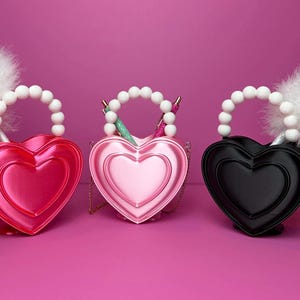 May include: Three heart-shaped handbags in red, pink, and black, each with a beaded handle and a fluffy white pom-pom. The bags have a layered heart design and a chain strap. The background is a solid pink.