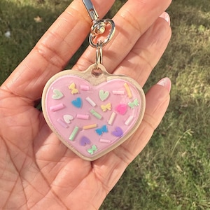 May include: A heart-shaped keychain with a pink background and colorful sprinkles and butterfly accents. The keychain has a gold-colored metal clasp and a clear border. The background is a grassy area.