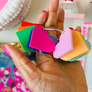Puede incluir: Un conjunto de etiquetas en forma de coraz&oacute;n de varios colores, unidas a un anillo plateado. Las etiquetas son de color rosa, naranja, amarillo, verde, azul y morado. La imagen est&aacute; bien iluminada, mostrando los colores vibrantes.