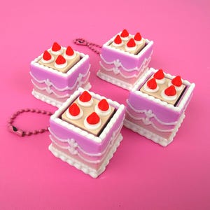 May include: Four miniature cake-shaped keychains. Each keychain features a square cake with pink and white frosting, a tan top, and red strawberry decorations. A pink beaded chain is attached to one keychain. The background is a solid pink.