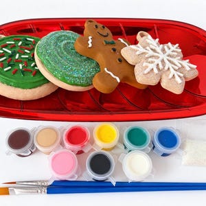 Puede incluir: Una bandeja roja con forma de hoja contiene cuatro adornos con forma de galleta: uno con glaseado verde y virutas, uno con purpurina verde, un hombre de jengibre marrón y un copo de nieve blanco. Incluye diez botes de pintura, dos pinceles y purpurina.