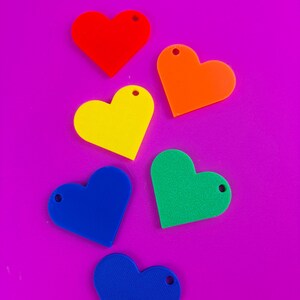 Puede incluir: Seis dijes en forma de coraz&oacute;n en rojo, naranja, amarillo, verde y dos tonos de azul. Cada dije tiene un peque&ntilde;o agujero en la parte superior. Dispuestos sobre un fondo morado brillante.