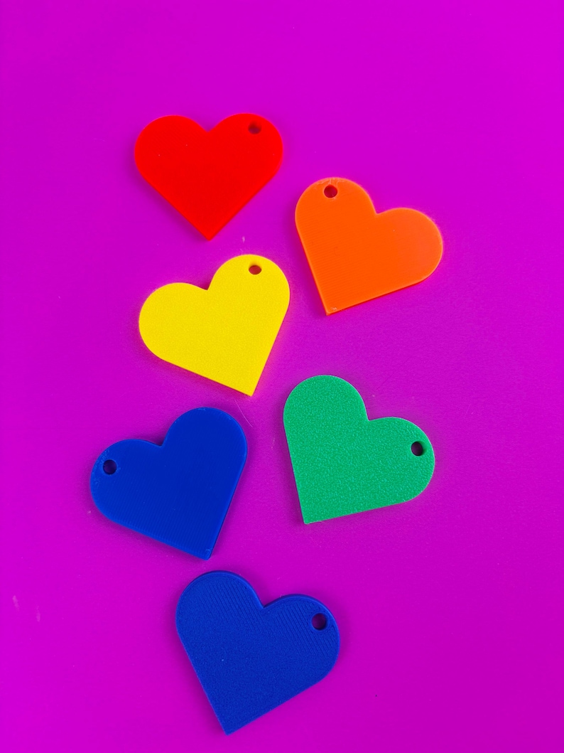 Puede incluir: Seis dijes en forma de coraz&oacute;n en rojo, naranja, amarillo, verde y dos tonos de azul. Cada dije tiene un peque&ntilde;o agujero en la parte superior. Dispuestos sobre un fondo morado brillante.