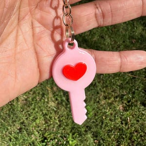 May include: A pink key-shaped keychain with a red heart in the center, attached to a gold chain. The keychain is held against a background of green grass.
