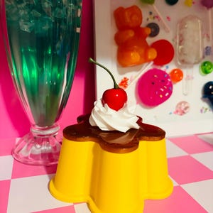 May include: A yellow, flower-shaped dessert with a dark brown topping, white whipped cream, and a red cherry. A tall glass of blue-green beverage with ice and a decorative frame with candy are in the background.