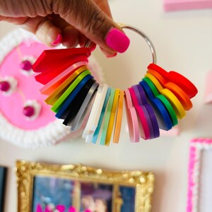 Puede incluir: Un anillo de muestras de colores del arco&iacute;ris, sostenido por una mano con esmalte de u&ntilde;as rosa. El anillo presenta una variedad de colores, incluyendo rojo, naranja, amarillo, verde, azul y morado. Las muestras est&aacute;n dispuestas en un patr&oacute;n circular.