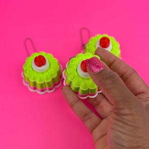 May include: Three lime green and white decorative ornaments with a red center and a silver loop for hanging. The ornaments are on a bright pink background. One ornament is being held by a hand with pink glitter nail polish.