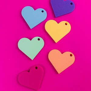 Puede incluir: Seis recortes de espuma en forma de coraz&oacute;n en azul, morado, amarillo, verde, naranja y rosa, cada uno con un peque&ntilde;o agujero en la parte superior. Los corazones est&aacute;n dispuestos sobre un fondo rosa brillante.