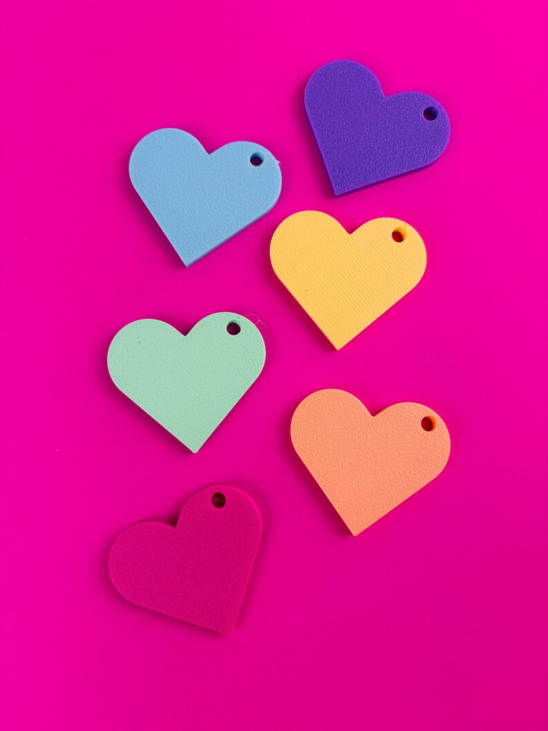 Puede incluir: Seis recortes de espuma en forma de coraz&oacute;n en azul, morado, amarillo, verde, naranja y rosa, cada uno con un peque&ntilde;o agujero en la parte superior. Los corazones est&aacute;n dispuestos sobre un fondo rosa brillante.