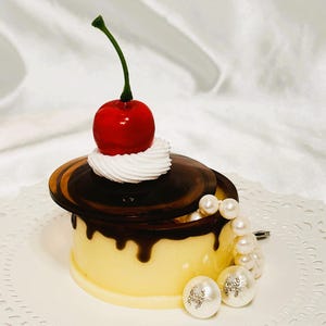Może przedstawiać: Biało-żółte ciasto z polewą czekoladową i czerwoną wiśnią na wierzchu. Ciasto znajduje się w przezroczystym pojemniku z pokrywką. Pojemnik stoi na białym obrusie.