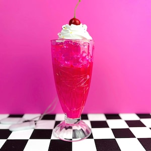 May include: A tall, clear glass filled with a bright pink liquid and ice cubes, topped with a generous swirl of white whipped cream and a single red cherry. The glass has a textured, faceted design and sits on a base, creating a classic soda fountain look.