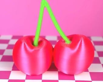 May include: Two bright red cherry sculptures with a vibrant green stem. The cherries are set against a pink checkered surface and a solid pink background. The image is a close-up shot, highlighting the smooth texture and color of the cherries.