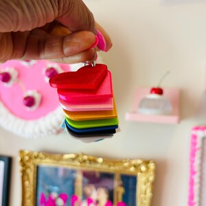 Puede incluir: Una pila de dijes acr&iacute;licos en forma de coraz&oacute;n de colores vibrantes: rojo, rosa, naranja, amarillo, verde, azul y negro. Los dijes son sostenidos por una mano con esmalte de u&ntilde;as rosa. El fondo incluye arte enmarcado y elementos decorativos.