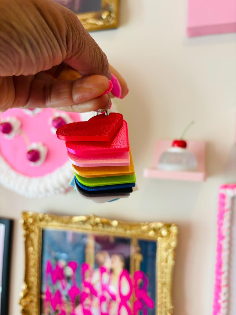 Puede incluir: Una pila de dijes acr&iacute;licos en forma de coraz&oacute;n de colores vibrantes: rojo, rosa, naranja, amarillo, verde, azul y negro. Los dijes son sostenidos por una mano con esmalte de u&ntilde;as rosa. El fondo incluye arte enmarcado y elementos decorativos.