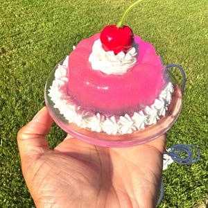 May include: A pink, gelatinous dessert with white frosting and a red cherry on top, presented on a clear plate. The dessert is held in a hand against a backdrop of green grass.