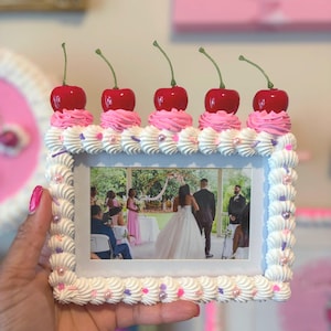 May include: A decorative photo frame with a cake-like design. The frame is white with piped frosting, pink and purple pearl accents, and topped with five red cherries. The frame holds a photo of a wedding ceremony.