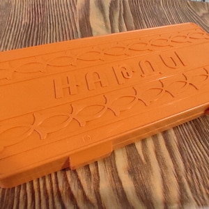 Puede incluir: Caja de plástico naranja con la palabra "HAWAII" en relieve en la parte superior. La caja tiene un patrón decorativo de círculos entrelazados.