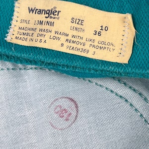 May include: A close-up of a teal blue Wrangler brand clothing label with white stitching. The label has the text "Wrangler", "STYLE 13MINM", "SIZE", "LENGTH", "10", "36", "MACHINE WASH WARM WITH LIKE COLOR", "TUMBLE DRY LOW, REMOVE PROMPTLY", "MADE IN USA", "B 9EAEH369 3".