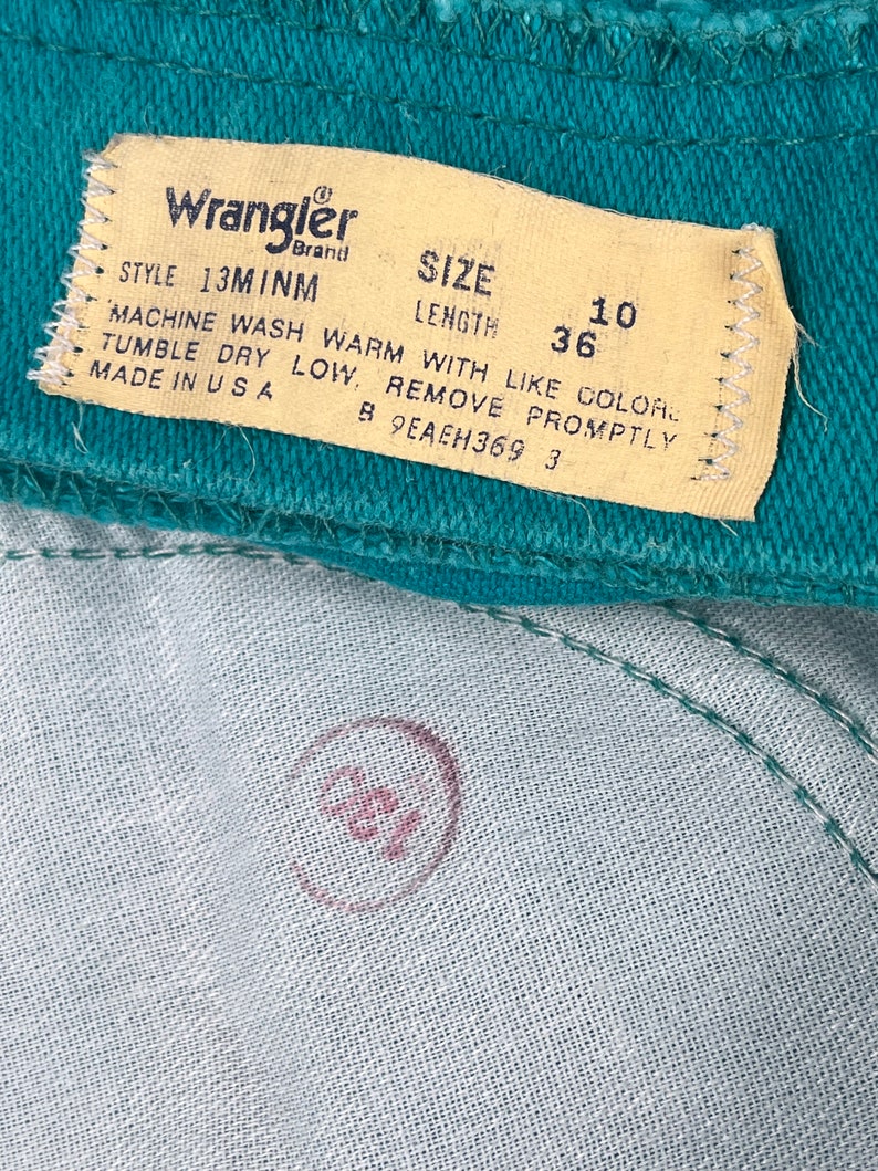 May include: A close-up of a teal blue Wrangler brand clothing label with white stitching. The label has the text "Wrangler", "STYLE 13MINM", "SIZE", "LENGTH", "10", "36", "MACHINE WASH WARM WITH LIKE COLOR", "TUMBLE DRY LOW, REMOVE PROMPTLY", "MADE IN USA", "B 9EAEH369 3".