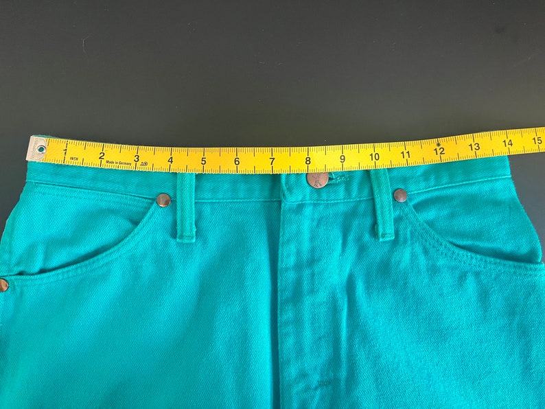May include: Teal blue denim jeans with a yellow measuring tape across the waistband. The tape reads 1 through 15.