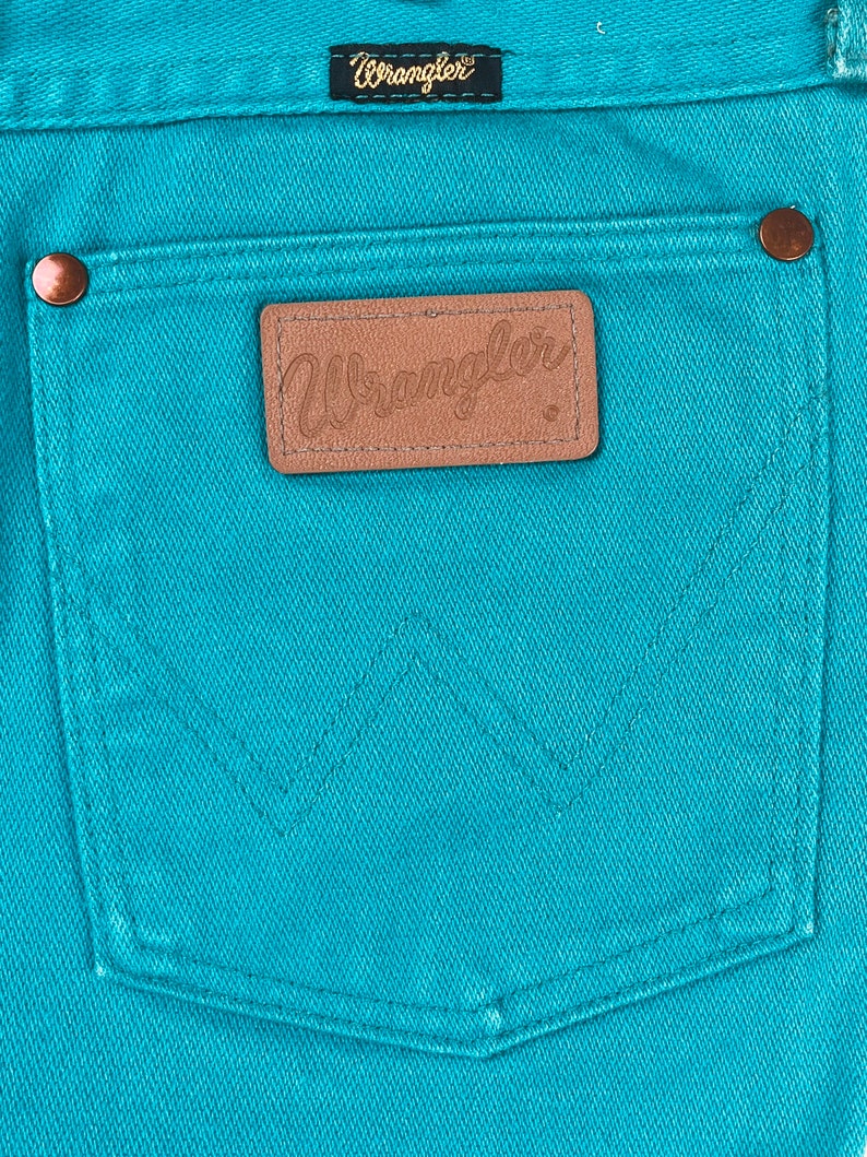 May include: A close-up of a turquoise blue pair of jeans with a brown leather patch that says "Wrangler" on the back pocket.