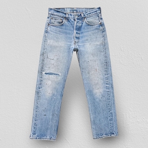 May include: Light blue denim jeans featuring a high-waisted fit, a button-fly closure, and a distressed finish with a horizontal tear on the right thigh. The fabric displays a faded wash with subtle whiskering and light wear throughout the legs.