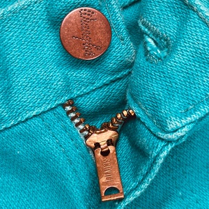 May include: Close-up of a turquoise blue denim zipper with a copper-colored zipper pull that reads "SCOVILL".