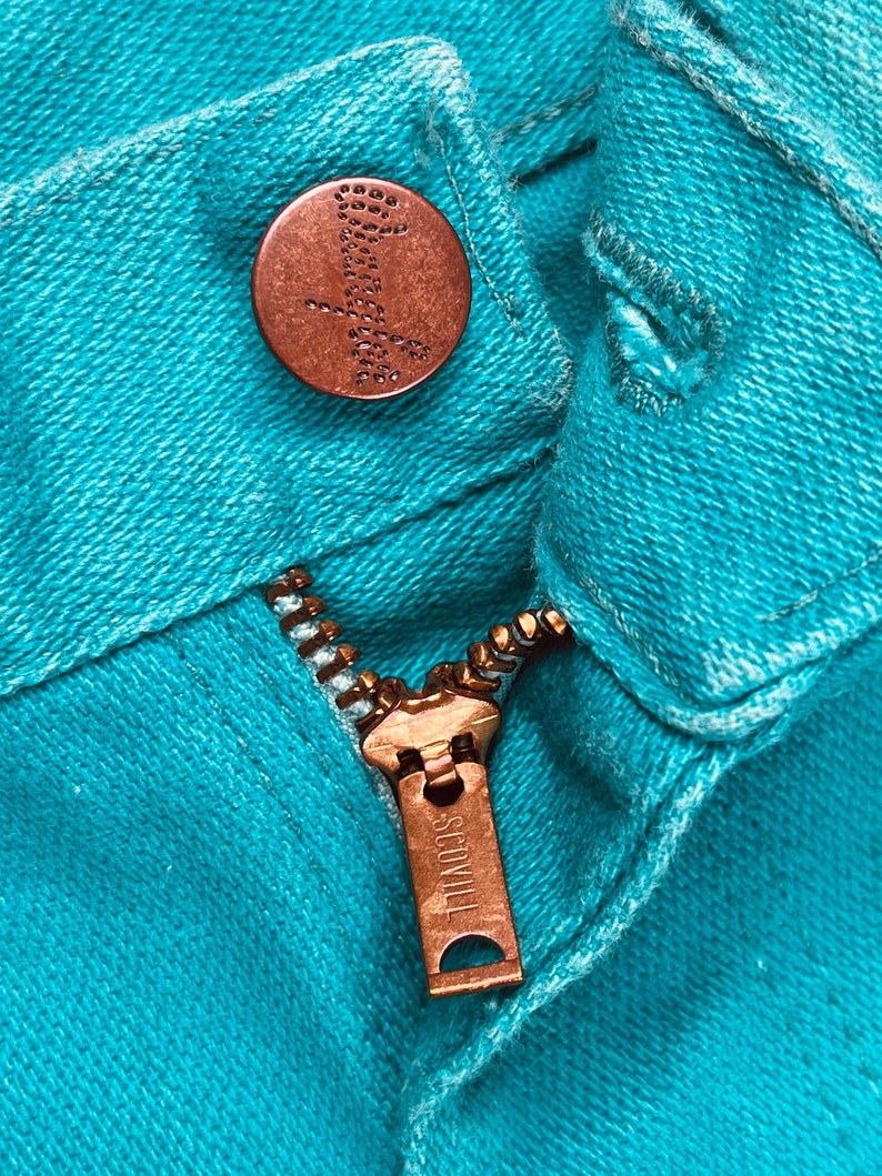 May include: Close-up of a turquoise blue denim zipper with a copper-colored zipper pull that reads "SCOVILL".